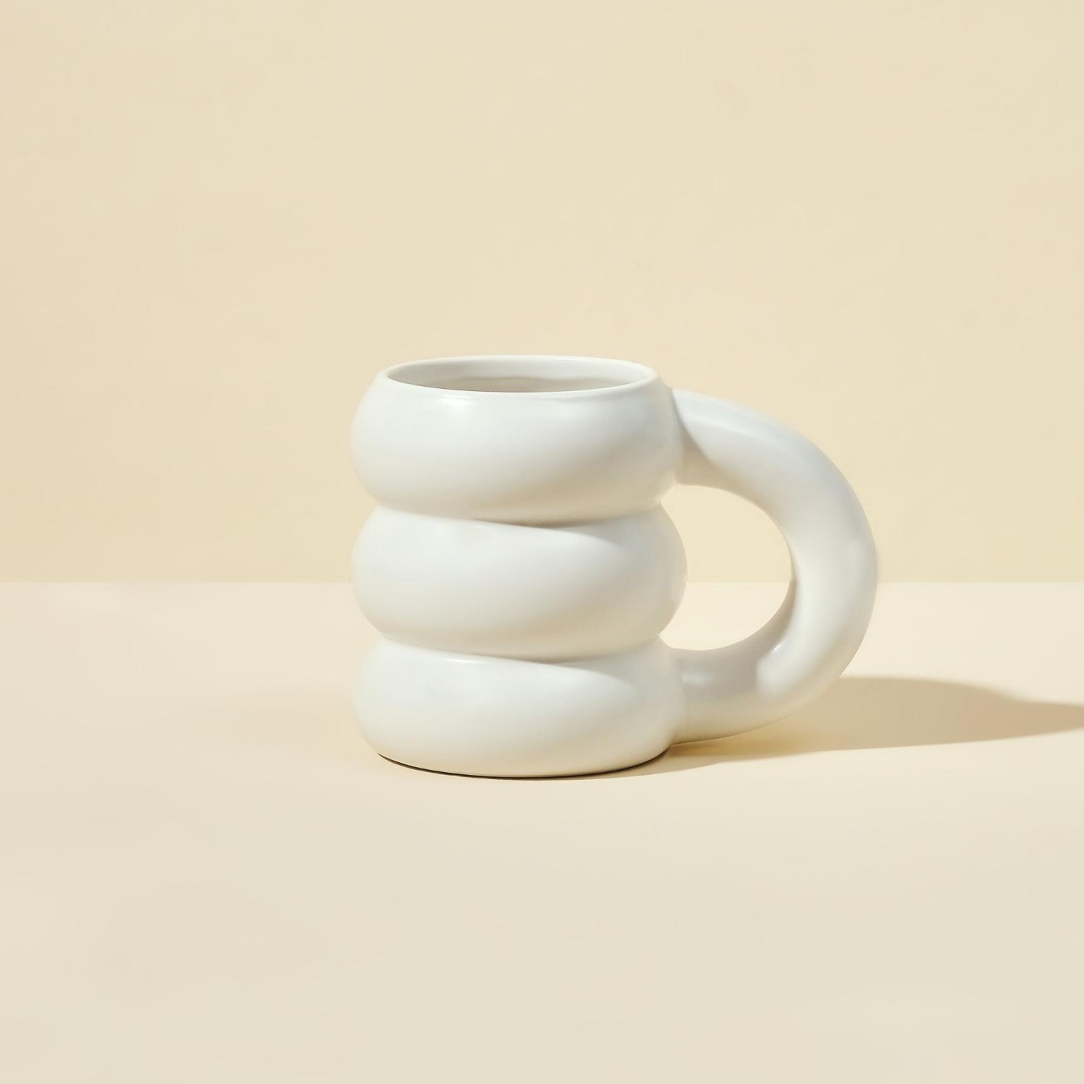 The limited edition Cloud Mug by Blume, featuring a white ceramic body with a unique spiral design and oversized handle, rests on a light beige surface against a matching backdrop.
